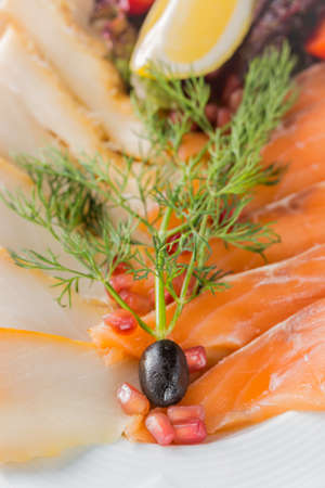 Salmon fillet pieces, sturgeon pieces served with lemon, black olives, herbs, cherry tomato and pomegranate seeds on white plate close up with selective focus.の写真素材