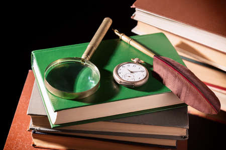 Literature concept. Old books with feather pen, magnifying glass and old vintage clock on black...