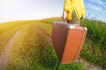 Young woman carries brown vintage suitcase in the field road during summer sunset. Travel conceptの写真素材
