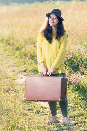 Beautiful happy smiling young woman with brown vintage suitcase and black hat in the field roadの写真素材