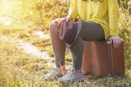 Tired girl sitting and resting on brown vintage suitcase in the field road during summer sunset. Toned image and travel concept.の写真素材