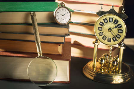 Literature concept. Magnifying glass near vintage clocks and old books against black background.の写真素材
