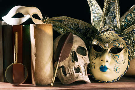 Theater and drama concept. Human skull, venetian masks with old scroll, books, magnifying glass and quill on wooden tableの写真素材