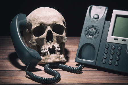 Office work concept. Human skull model with modern telephone on wooden table and black backgroundの写真素材