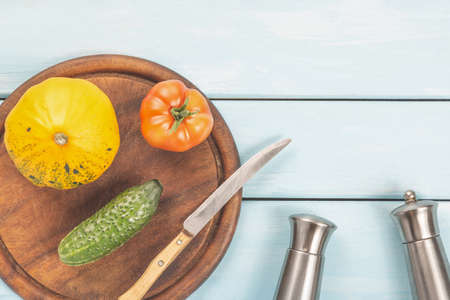 Salad delicatessen preparation. Vegetarian healthy food. Tomato, cucumber and bush pumpkin on cutting board with knife, saltcellar, pepper shaker on blue wooden backgroundの写真素材
