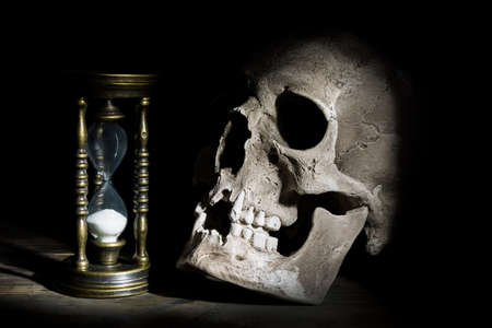 Skull and vintage hourglass on wooden background under beam of light.の写真素材