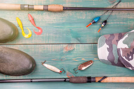 Fishing theme. Frame from fishing rods with fishing tackles, rubber boots, camouflage cap and fishing buoy on green wooden backgroundの写真素材
