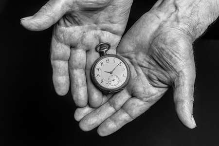 Time concept. Vintage clock in old senior wrinkled male hands on black background.の写真素材