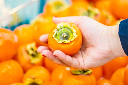 Woman holding orange date plum in the supermarket. Vegetables and fruits in the background. Shopping...