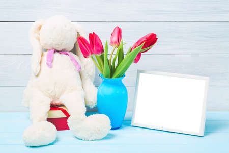 Colorful red spring tulip flowers in nice blue vase, blank photo frame and stuffed toy bunny on light wooden background as greeting card. Mothersday or spring concept.の写真素材
