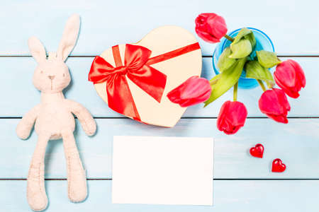 Colorful red spring tulip flowers in nice blue vase, blank photo frame and stuffed toy bunny with decorative heart on light wooden background as greeting card. Mothersday or spring concept. Top view.の写真素材