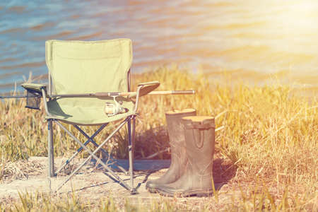Fishing concept. Fishing chair on green grass with boots and spinning rod near lake. Toned.の写真素材