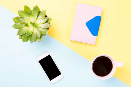 Conceptual workspace or business concept. Green plant in a pot, cup of coffee, notebook and modern mobile phone on blue and yellow pastel background. Top view.の写真素材
