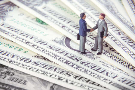 Business deal or agreement and success concept. Two miniature businessmen shaking hands while standing on american dollar money backgroundの写真素材
