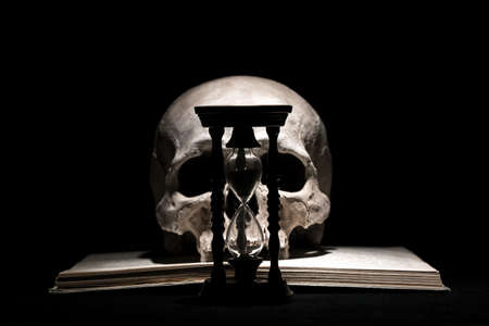 Human skull on old open book with vintage hourglass on black background. Drama and time conceptの写真素材