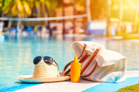 Straw hat with sunglasses near sunscreen lotion bottle and beach bag against the surface of blue...