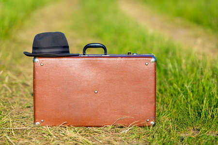 Travel and adventure concept. Vintage brown suitcase and hat on the road in the green fieldの写真素材