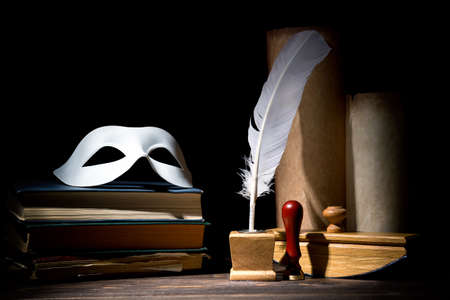 Drama or theater and literature concept. Old inkstand with feather near scrolls with mask on books against black background. Dramatic light.の写真素材