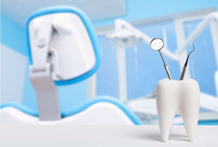 Oral dental hygiene. Healthy white tooth and dentist mirror with explorer probe instrument against blurred dentist Office background with dental chair and lampの写真素材
