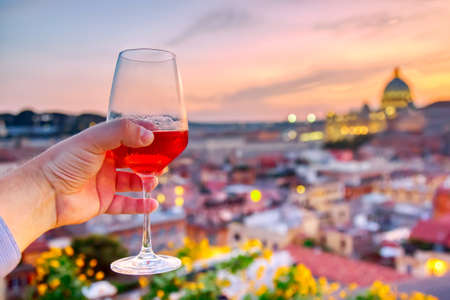 Male hand holding red wine glass against the aerial cityscape view of Rome at sunset with St Peter Cathedral in Vaticanの写真素材