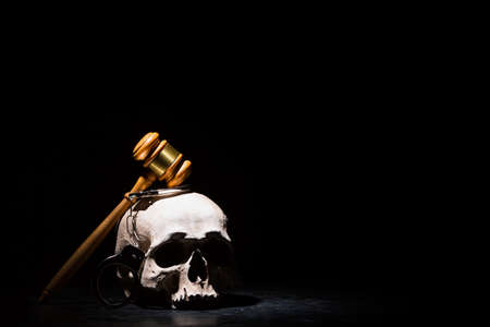 Legal law, justice and murderment concept. Wooden judge gavel hammer on human skull with handcuffs against black background. Free spaceの写真素材