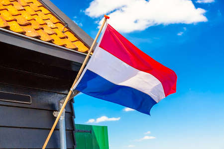 Flag of the Netherlands waving in the wind against blue sky and old dutch house in Zaanse Schans village.の写真素材