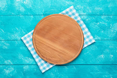 Round wooden cutting board for pizza with blue plaid tablecloth on blue wooden table. Top view. Mockup for food projectの写真素材