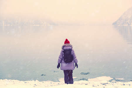 Hiker girl with backpack in down jacket and red cap standing near lake in winter. Travel conceptの写真素材