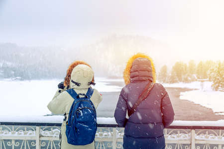 Hiker girls in down jackets standing near river against winter forest and mountains. Travel conceptの写真素材