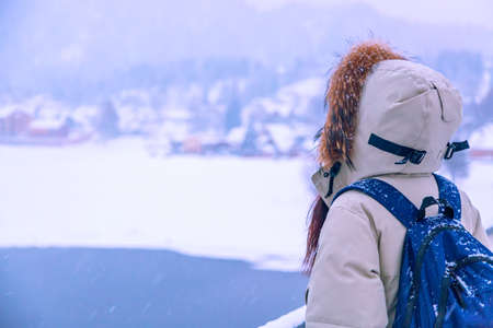 Hiker girl with backpack in down jacket and hood standing near lake in winter. Travel conceptの写真素材