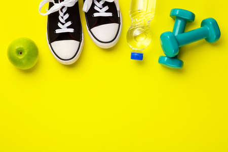 Fitness and sport concept. Sneakers, green apple. dumbbells and bottle of water on bright yellow background. Free space.の写真素材