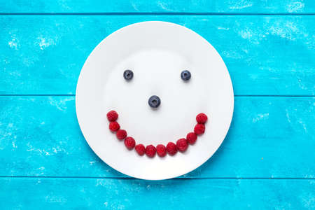 Smiling face from blueberries and raspberries on white plate and blue wooden table. Diet and healthy...
