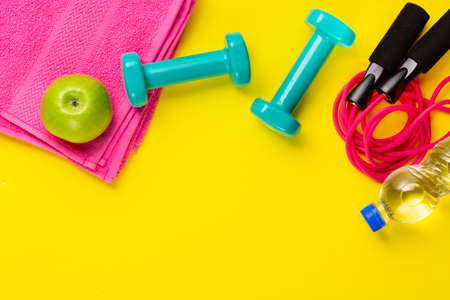 Fitness and sport concept. Skipping rope, green apple, dumbbells, bottle of water and pink towel on bright yellow background. Free space.の写真素材