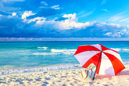 Colorful red and white umbrella with beach bag on the ocean beach against beautiful blue sky and clouds. Relaxation, vacation idyllic backgroundの写真素材