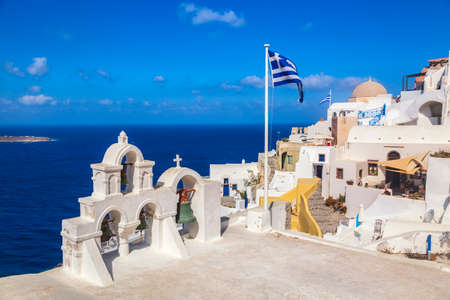 Greek orthodox church with bells and greek flag against famous white houses on Santorini island, Aegean sea, Greeceの写真素材