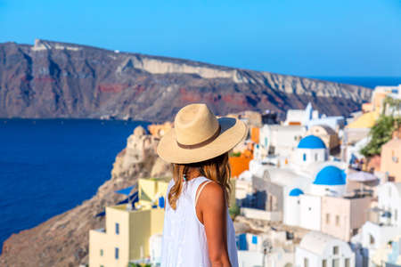 Beautiful tourist girl in straw hat against Oia village with famous white houses and blue churches on Santorini island, Aegean sea, Greece. Travel concept.の写真素材