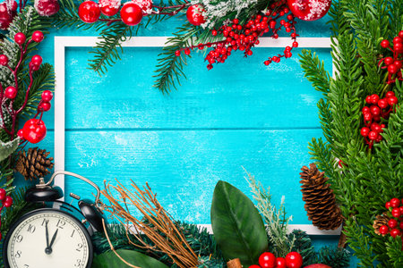 Blank white photo frame, vintage clock, fir tree branches, red berries and decorations on blue wooden table. Winter Christmas celebration concept. Free space for your text.の写真素材
