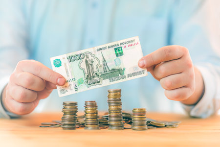 Saving money. Accounting, business, budget and finance concept. Male hands holding one thousand Russian roubles banknote above coins stacksの写真素材