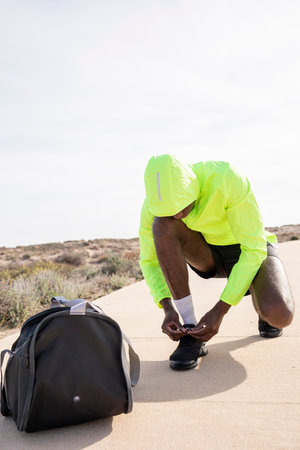 Vertical Shoot of Young black runner with bag and yellow rain jacket ties his laces and gets ready...