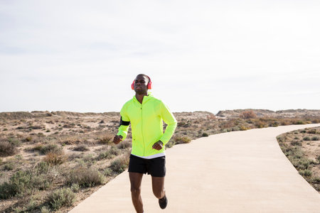 Young smiling black runner in a yellow rain jacket who enjoys listening to music on his cell phone...