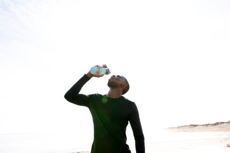 Young black sportsman on the beach who opens a bottle of water with his hands while resting.の写真素材