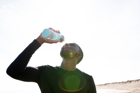 Young black sportsman on the beach drinking water from a bottle while resting. Flare that goes through the young man's faceの写真素材
