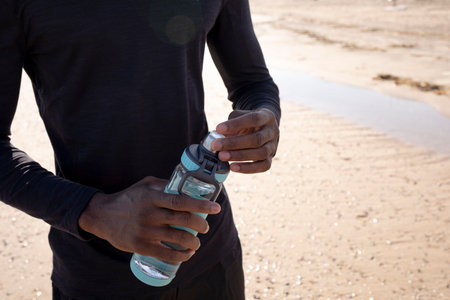 Young black sportsman on the beach who opens a bottle of water with his hands while resting.の写真素材