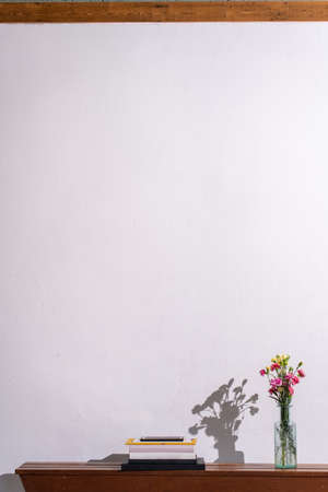 Pile of books and a vase with flowers on a wooden shelf against a white wall