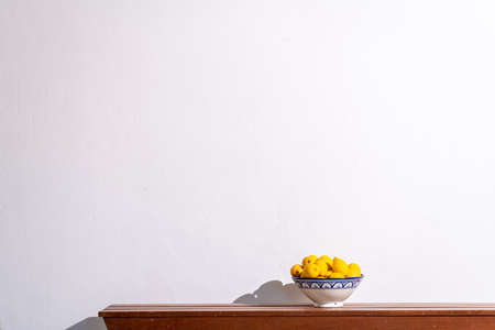 Yellow lemons in a ceramic bowl on a wooden table against a white wall. Copy