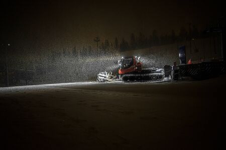Snowcat with front lights preparing a slope at night in mountains at skiing resort with the snowfallの写真素材