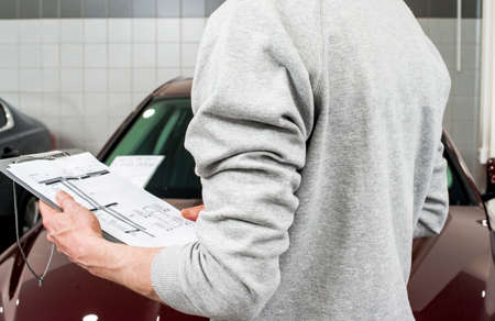 car service, repair, maintenance concept - mechanic writing to the clipboard at workshop warehouse, technician doing the checklist for repair machine a car in the service, detailingの写真素材