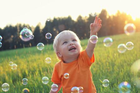 Cute toddler blond boy playing with soap bubbles on summer field. Happy child summertime concept. Authentic lifestyle image.の写真素材