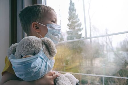 Sad illness child on home quarantine. Boy and his teddy bear both in protective medical masks sits on windowsill and looks out window. Virus protection, coronavirus pandemic, prevention epidemic.の写真素材