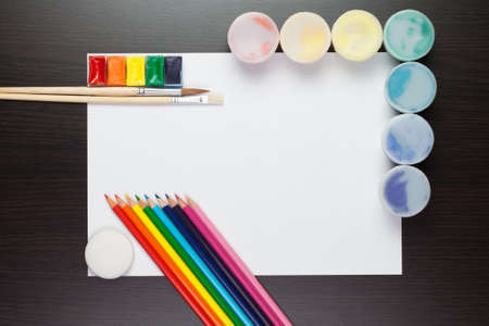 blank sheet of paper on the brown table with colorful paints and pencilsの写真素材